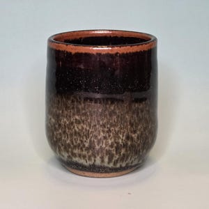 May include: A cylindrical ceramic cup featuring a glossy dark brown glaze on the upper portion that transitions into a mottled, textured beige and brown finish toward the base. The rim is a solid, smooth dark brown with a visible terracotta-colored edge.