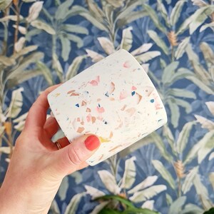 May include: A white terrazzo planter with pink, blue and brown flecks. The planter is being held in a hand with red nail polish.