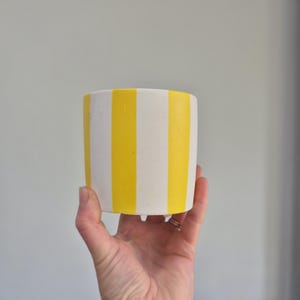 May include: A small, cylindrical planter with vertical yellow and white stripes. The planter is held in a hand. It has three small feet. The background is a plain, light grey colour. The planter is suitable for small plants.