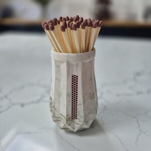 May include: A ceramic match holder with a geometric design, filled with wooden matches. The holder is off-white with a marbled effect and a vertical strip of dark red dots. The match heads are brown.