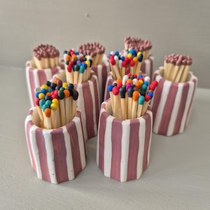 May include: A selection of match holders, each filled with matches. The holders are white with pink stripes. Some matches have brown tips, others have colourful tips in a variety of shades.