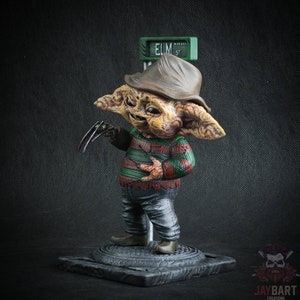 May include: A small, detailed figurine of Freddy Krueger from the Nightmare on Elm Street movie franchise. The figurine is painted in a realistic style and features Freddy's signature red and green striped sweater, brown hat, and clawed glove. The figurine is standing on a base with a street sign that reads "ELM ST."