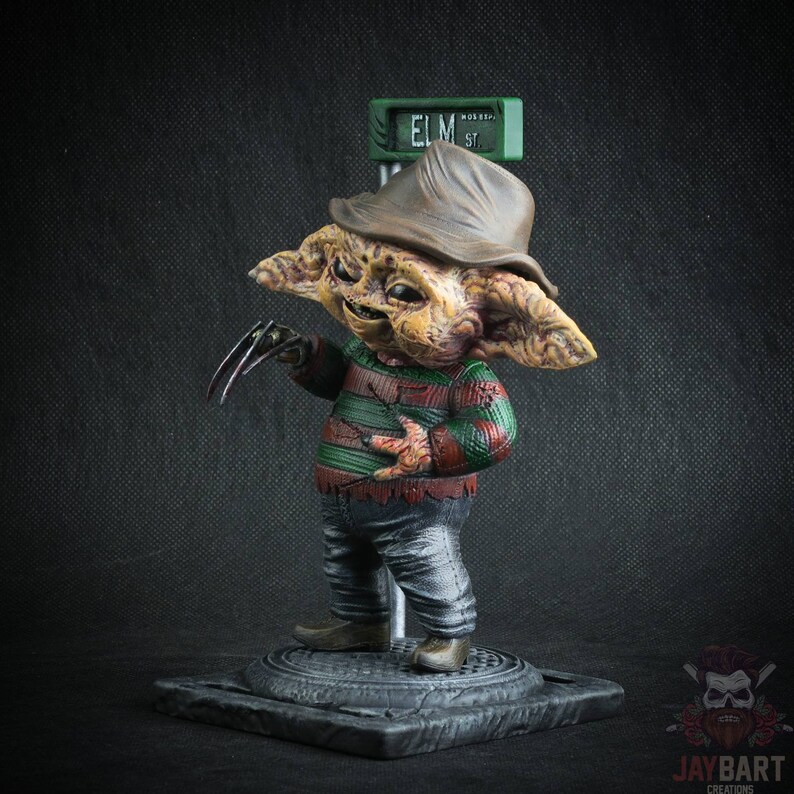 May include: A small, detailed figurine of Freddy Krueger from the Nightmare on Elm Street movie franchise. The figurine is painted in a realistic style and features Freddy's signature red and green striped sweater, brown hat, and clawed glove. The figurine is standing on a base with a street sign that reads "ELM ST."
