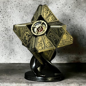 May include: A gold-colored, geometrically shaped, decorative object with intricate details. It is mounted on a black stand with a swirling design.