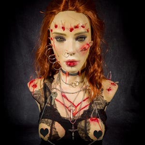 Puede incluir: Un busto de maniquí con cabello largo color caoba, textura de piel realista, múltiples piercings faciales y en el pecho, y fragmentos de vidrio incrustados con acentos de pintura roja. Viste un top de encaje negro, cadenas plateadas y una cruz.