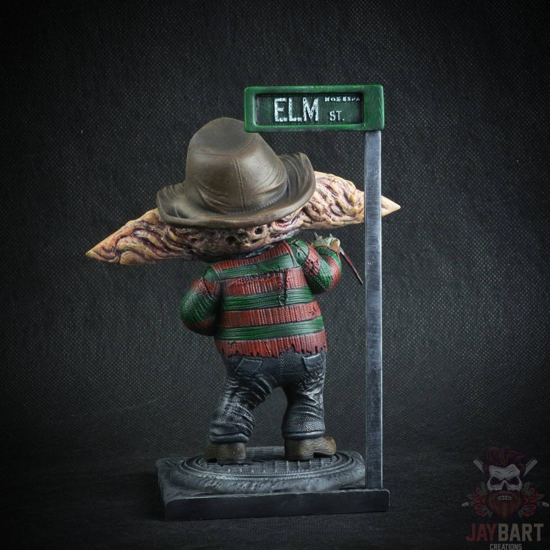 May include: A Freddy Krueger figurine with a red and green striped sweater, brown hat, and a glove with a razor blade. The figurine is standing on a base with a street sign that reads "ELM ST."