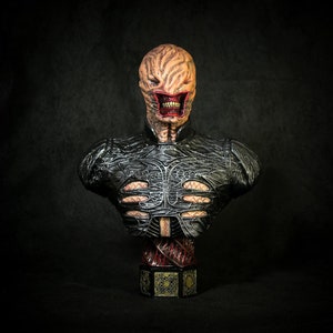 May include: A detailed bust of a character with a textured, exposed brain-like head and a wide, open mouth revealing teeth. The figure has a dark, textured upper body with intricate patterns and a decorative base. The color palette includes shades of brown, red, and black.