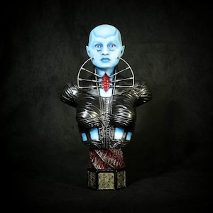 May include: A blue-skinned bust sculpture with a detailed black leather-like jacket and a red accent on the chest. The sculpture has a silver collar and is mounted on a decorative base with gold and red accents. The bust has a serene expression.