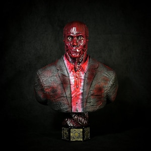 May include: A red and black bust sculpture of a figure with exposed facial muscles, wearing a gray blazer over a red collared shirt. The sculpture is mounted on a decorative, geometric base with gold accents. The background is black.
