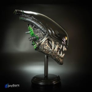 May include: A detailed sculpture of an Alien Xenomorph head, featuring a glossy black exterior and intricate internal detailing. The sculpture is mounted on a black stand, showcasing the creature's sharp teeth and biomechanical design. Green accents highlight the alien's features.