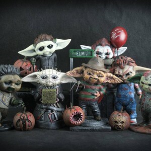 May include: A collection of eight horror movie character figurines, each with a unique design and paint scheme. The figurines are all in the style of the popular "Baby Yoda" character, with large ears and big eyes. The characters include Freddy Krueger, Jason Voorhees, Chucky, Pennywise, Leatherface, Michael Myers, and two others. The figurines are all standing on a black surface with a few pumpkins in front of them.