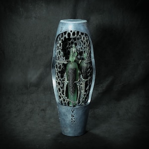 May include: A silver-coloured, cylindrical vase with a textured, honeycomb-like design. The vase is filled with green, translucent, organic-looking forms.