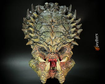 Könnte beinhalten: Eine detaillierte Predator-Maske mit texturierter, reptilienartiger Erscheinung. Die Maske hat scharfe, hervorstehende Stacheln, große Stoßzähne und einen bedrohlichen, offenen Mund. Die Farbpalette umfasst Grün-, Braun- und Beigetöne mit roten Akzenten. Der Text "JayBart" ist sichtbar.