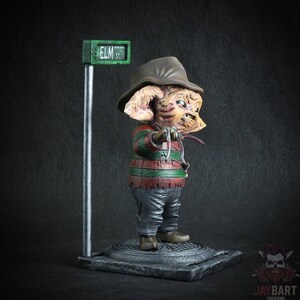 May include: A small, detailed figurine of Freddy Krueger from the Nightmare on Elm Street movie franchise. The figurine is wearing a striped sweater, brown hat, and gloves with blades. The figurine is standing on a base with a street sign that reads "ELM ST."