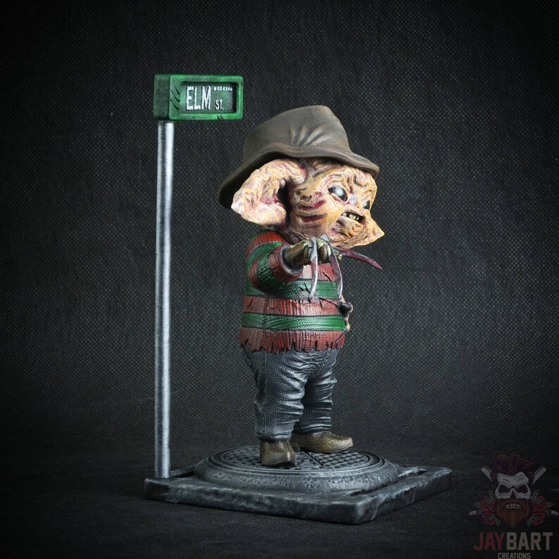 May include: A small, detailed figurine of Freddy Krueger from the Nightmare on Elm Street movie franchise. The figurine is wearing a striped sweater, brown hat, and gloves with blades. The figurine is standing on a base with a street sign that reads "ELM ST."