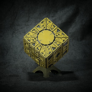 May include: A gold-colored metal puzzle box with intricate designs and a black base. The box is on a black fabric surface.