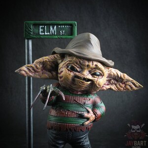 May include: A small, detailed figurine of a fictional character with a brown hat, green and red striped shirt, and a menacing expression. The character is holding a pair of sharp, metal claws. A green street sign with the text "ELM ST." is visible in the background.