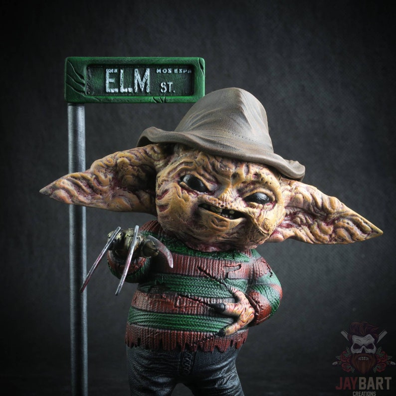 May include: A small, detailed figurine of a fictional character with a brown hat, green and red striped shirt, and a menacing expression. The character is holding a pair of sharp, metal claws. A green street sign with the text "ELM ST." is visible in the background.