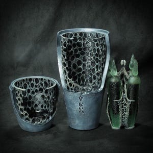 May include: Three decorative vases with a dark, textured interior and a metallic gray exterior. The vases have a unique, organic design with a pattern of circular openings. One vase has a green, ornate top.