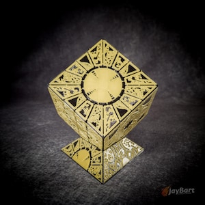 May include: A gold and black cube puzzle box featuring intricate etched geometric patterns and a central circular medallion on each face. The object is balanced on one corner, displaying detailed engravings and the text JayBart Creations.