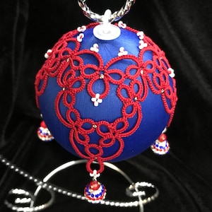 May include: A blue and red Christmas ornament with a red crocheted design and white beads. The ornament is on a silver stand.