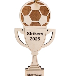 May include: A wooden soccer trophy ornament with a soccer ball design at the top. The trophy has the words "Strikers 2025" and "Matthew" engraved on it. A twine loop is attached for hanging.