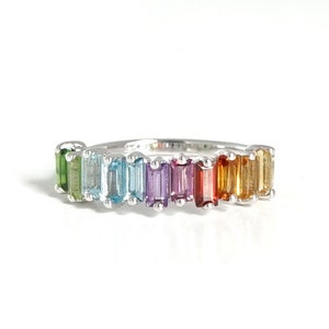 May include: A silver band ring featuring a rainbow of gemstones. The gemstones are set in a row and are emerald green, light blue, sky blue, purple, red, orange, and yellow.
