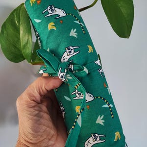 Könnte beinhalten: Ein aufgerolltes grünes Stofftuch mit einem sich wiederholenden Muster aus weißen Cartoon-Tieren mit gestreiften Schwänzen, Bananen und Blättern. Die Wickel ist mit einer passenden Stoffschleife gebunden. Es wird von einer Hand gehalten.