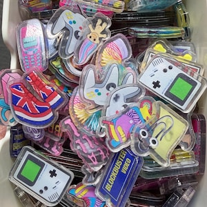 May include: A collection of assorted acrylic charms featuring nostalgic 90s imagery, including handheld gaming consoles, a Blockbuster Video sign, a paperclip character, a troll doll, a mobile phone, and platform sneakers with a Union Jack pattern.