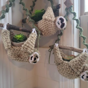 May include: Three crocheted sloth planters hanging from a branch. Each sloth is holding a small green succulent plant in a pot. The sloths are beige with brown faces and white accents.