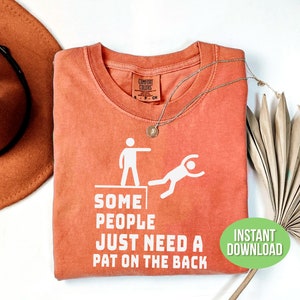 Pu&ograve; includere: Una maglietta color corallo con la scritta "SOME PEOPLE JUST NEED A PAT ON THE BACK" e una grafica di una persona che ne spinge un'altra da un bordo. Sono visibili una collana d'argento e un cappello.