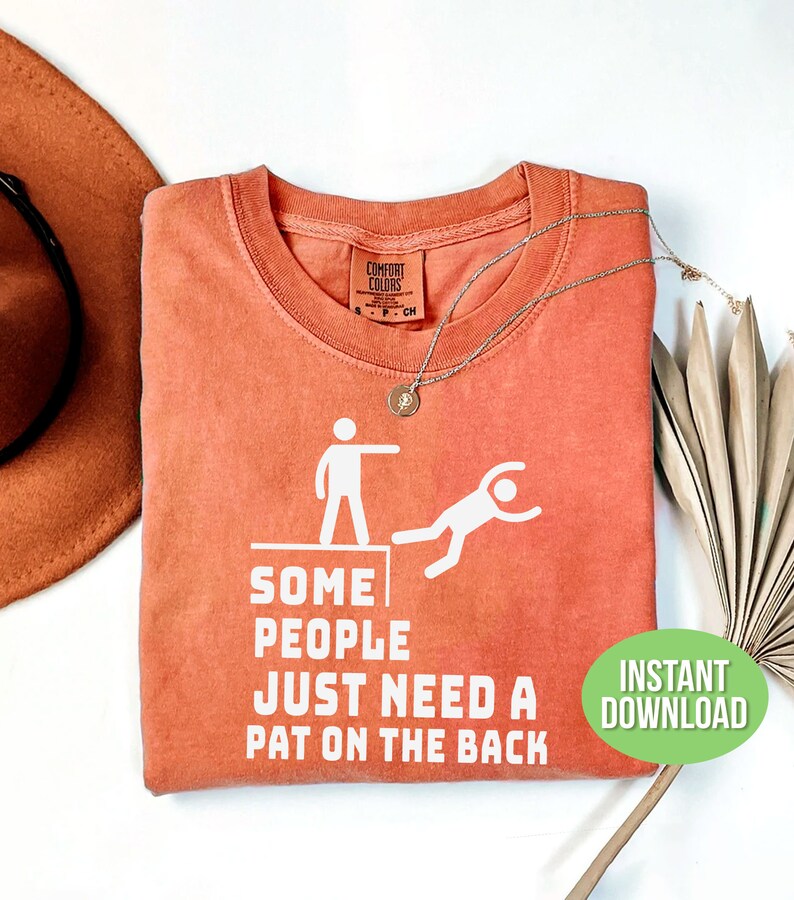 Pu&ograve; includere: Una maglietta color corallo con la scritta "SOME PEOPLE JUST NEED A PAT ON THE BACK" e una grafica di una persona che ne spinge un'altra da un bordo. Sono visibili una collana d'argento e un cappello.