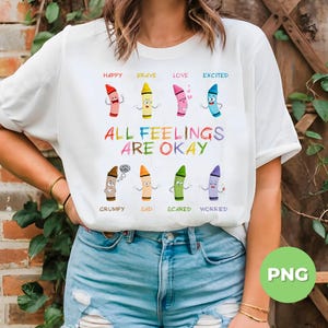Può includere: T-shirt bianca con grafica a pastelli colorati. La maglietta presenta la scritta "ALL FEELINGS ARE OKAY" e illustrazioni di pastelli che rappresentano diverse emozioni: felice, coraggioso, amore, eccitato, scontroso, triste, spaventato e preoccupato.