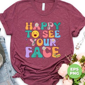 May include: A maroon t-shirt with the text "HAPPY TO SEE YOUR FACE" in colorful, retro-style letters. The text is decorated with flowers, stars, and vines. The shirt is photographed on a white background.