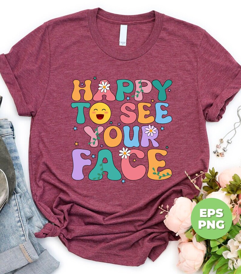 May include: A maroon t-shirt with the text "HAPPY TO SEE YOUR FACE" in colorful, retro-style letters. The text is decorated with flowers, stars, and vines. The shirt is photographed on a white background.