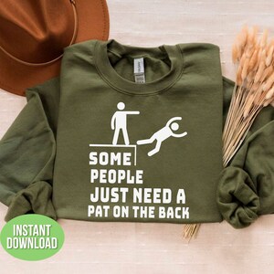 Pu&ograve; includere: Una felpa verde oliva con testo e grafica bianchi. Il testo recita "SOME PEOPLE JUST NEED A PAT ON THE BACK." Una grafica bianca raffigura una persona che ne spinge un'altra da una sporgenza. Le parole "INSTANT DOWNLOAD" sono in un cerchio verde.