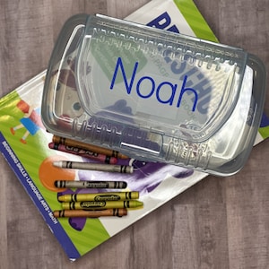 May include: A clear plastic pencil case with a blue label that says "Noah". The case is sitting on top of a children's book with colorful crayons inside.