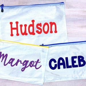 May include: Three clear plastic zippered bags with names written on them. The top bag has the name "Hudson" in red letters. The middle bag has the name "Margot" in purple letters. The bottom bag has the name "Caleb" in blue letters.