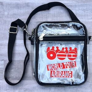May include: A clear crossbody bag with a black strap and trim. The bag features a red graphic and the words "WORLD TOUR ARIRANG" in red. The bag has a zipper closure and a silver-colored metal clasp.