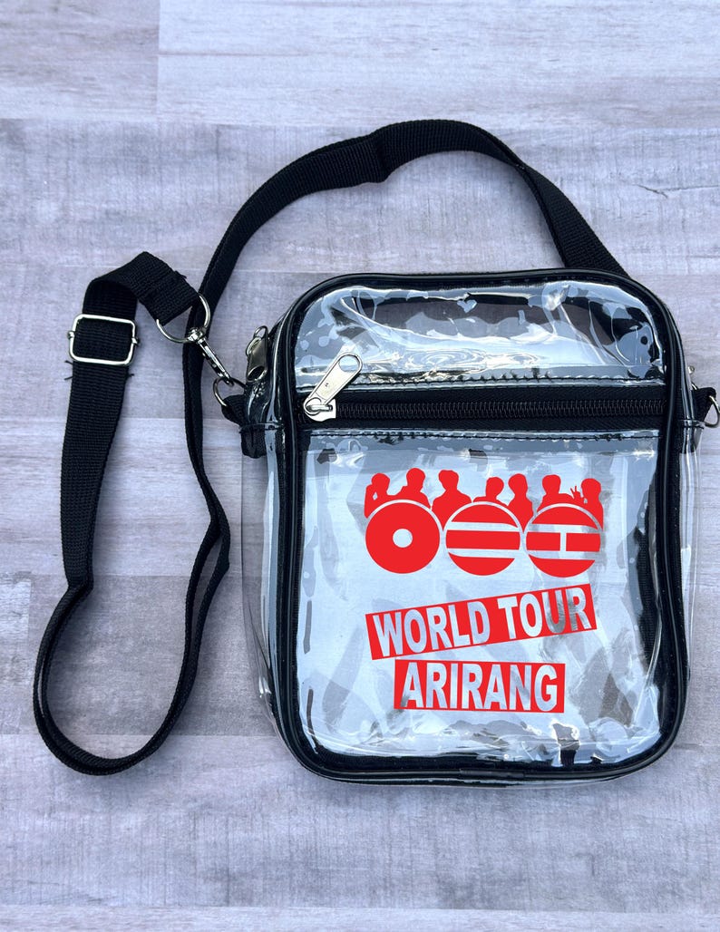 May include: A clear crossbody bag with a black strap and trim. The bag features a red graphic and the words "WORLD TOUR ARIRANG" in red. The bag has a zipper closure and a silver-colored metal clasp.