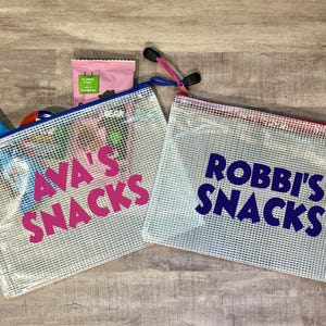 May include: Two clear mesh zippered bags with personalized text. The bag on the left says "AVA'S SNACKS" in pink letters. The bag on the right says "ROBBi'S SNACKS" in blue letters.
