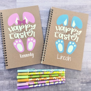 May include: Two brown spiral notebooks with personalized names "Kennedy" and "Lincoln". Each notebook features a bunny ear design with the text "Happy Easter" in white. The notebooks are accompanied by a stack of six colorful pens with Easter-themed designs.