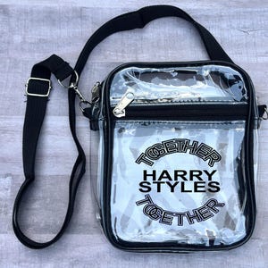 May include: Clear crossbody bag with black trim and a black adjustable strap. The bag features the text "TOGETHER HARRY STYLES TOGETHER" in black. The bag has a silver zipper and hardware.