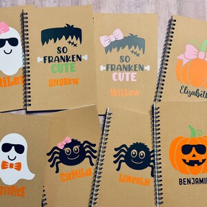 Puede incluir: Una colecci&oacute;n de cuadernos con espiral con dise&ntilde;os de Halloween. Cada cuaderno presenta un gr&aacute;fico diferente, incluyendo un fantasma, Frankenstein, una ara&ntilde;a y una calabaza, con nombres personalizados en naranja o negro.