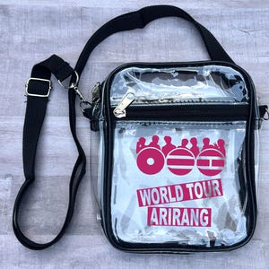 May include: Clear crossbody bag with a black strap and trim. The bag features a pink graphic with the words "WORLD TOUR ARIRANG" in white. The bag has a zipper closure and a silver-colored metal clasp.