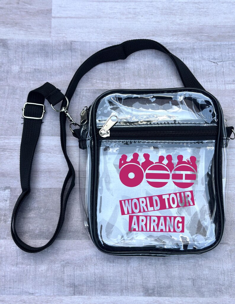May include: Clear crossbody bag with a black strap and trim. The bag features a pink graphic with the words "WORLD TOUR ARIRANG" in white. The bag has a zipper closure and a silver-colored metal clasp.