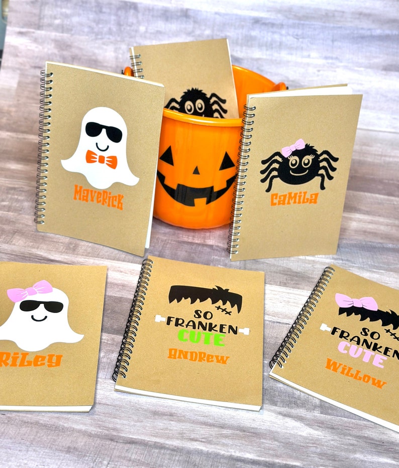Puede incluir: Una colecci&oacute;n de cuadernos con espiral con dise&ntilde;os de Halloween. Los cuadernos presentan ilustraciones de fantasmas, ara&ntilde;as y el monstruo de Frankenstein, junto con nombres como Maverick, Camila, Riley, Andrew y Willow. Tambi&eacute;n hay un cubo de calabaza naranja.