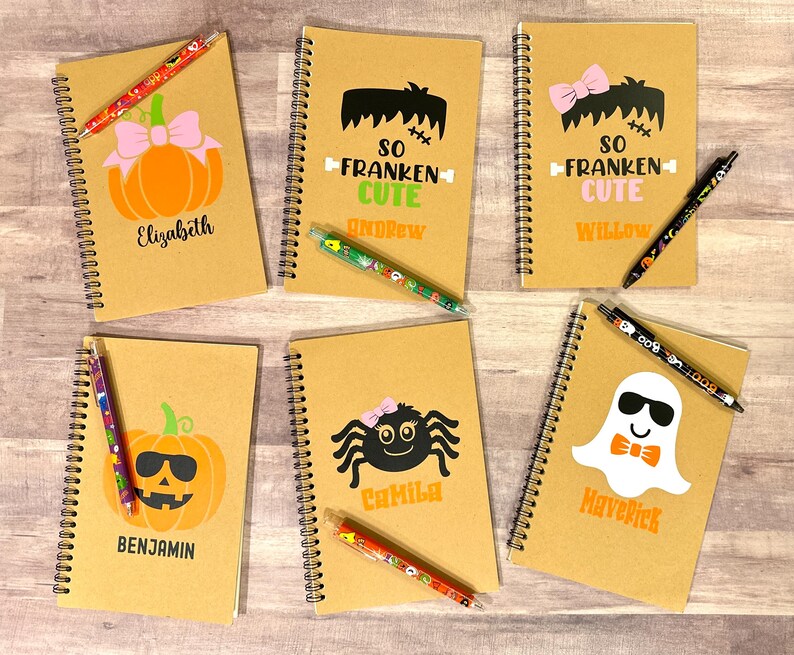 Puede incluir: Una colecci&oacute;n de cuadernos con espiral con dise&ntilde;os de Halloween. Cada cuaderno presenta un gr&aacute;fico diferente, incluyendo calabazas, un fantasma y una ara&ntilde;a, con nombres impresos debajo. Hay bol&iacute;grafos colocados encima de los cuadernos.