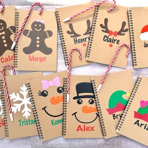 May include: A collection of spiral-bound notebooks with festive Christmas designs. Each notebook features a unique holiday character, including gingerbread people, reindeer, snowmen, and elves, with personalized names. Candy canes are placed on top.