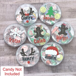 May include: Seven clear plastic containers filled with assorted candies. Each container has a different Christmas-themed design and a name printed on the front: Claire, Emerson, Caleb, Margot, Tristan, Henry, and Jacqueline.